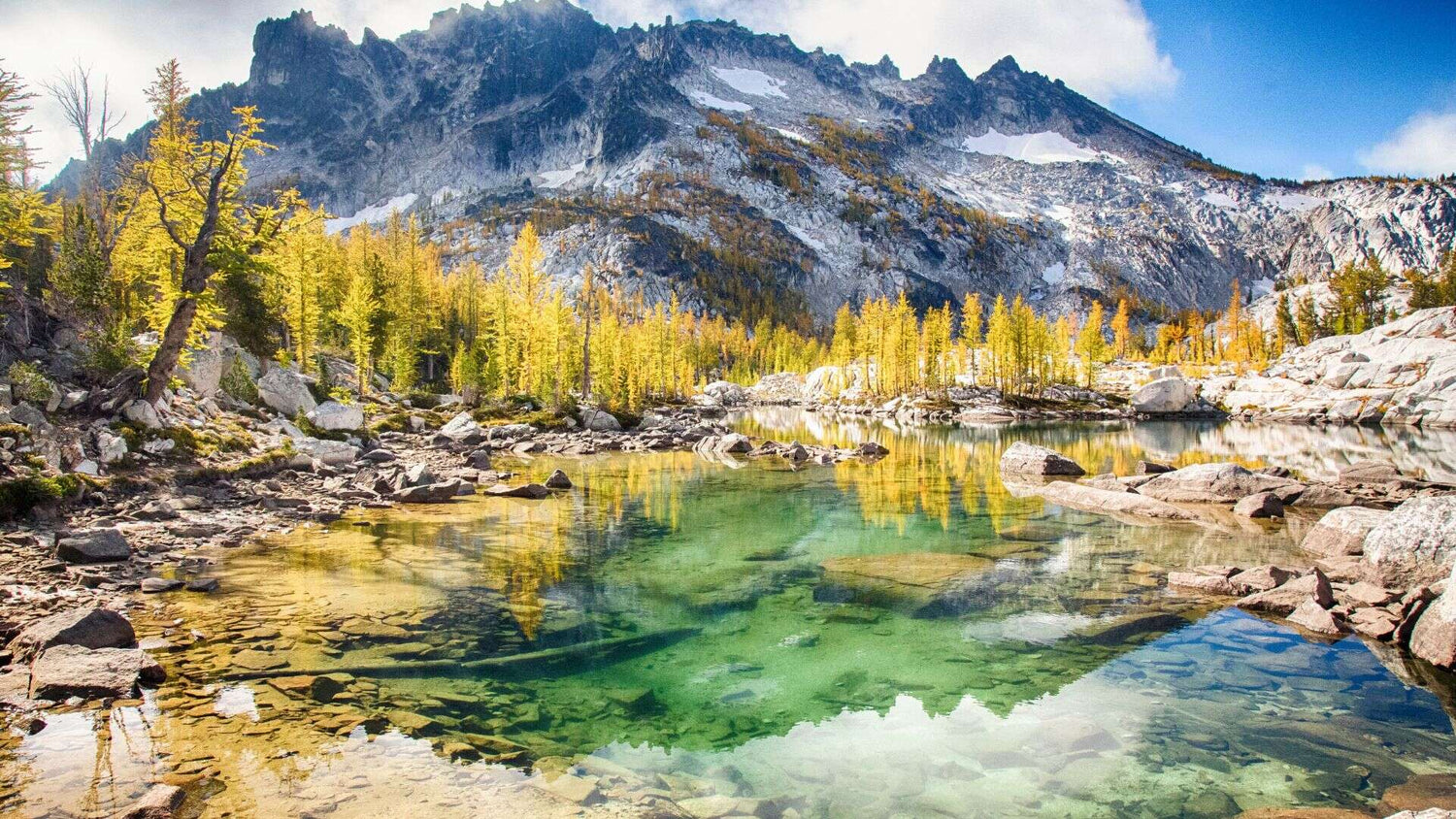 A view of The Enchantments in Washington features a clear pond in the middle and a mountain range in the background. - History By Mail